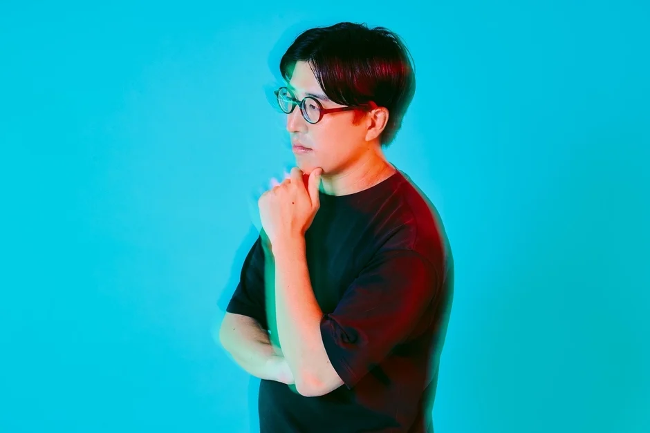 青い背景の前で、黒いTシャツと眼鏡をかけた男性