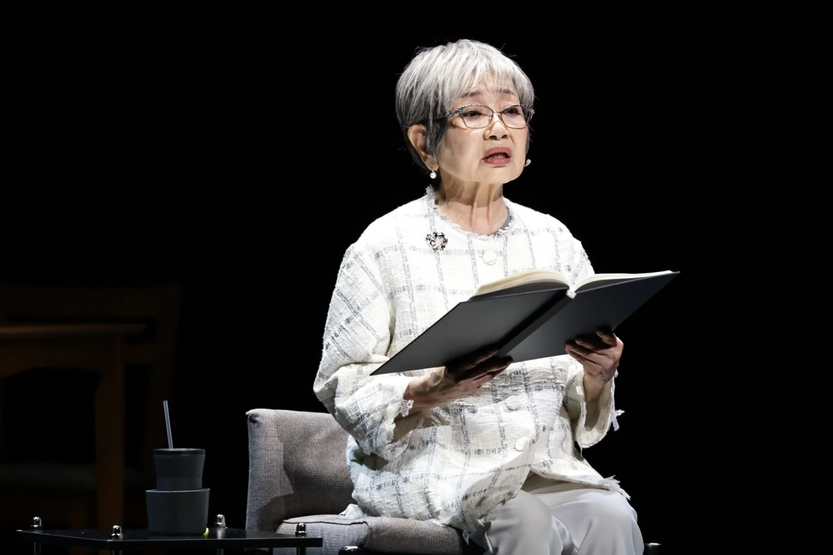 黒い背景のステージで、白髪と眼鏡の高齢女性が椅子に座り、本を開いて朗読している様子