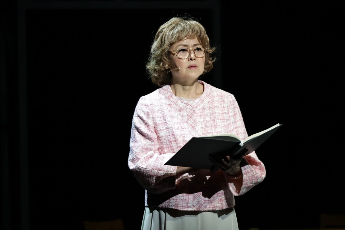 暗い舞台上で、眼鏡をかけた中年の女性がピンクのツイードジャケットを着用し、開いた本を手に何かを語っている様子
