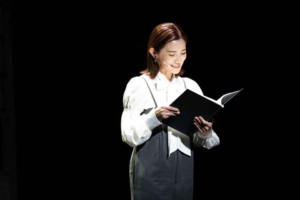 暗い背景の中、スポットライトを浴びた女性が笑顔で黒い本を読んでいます