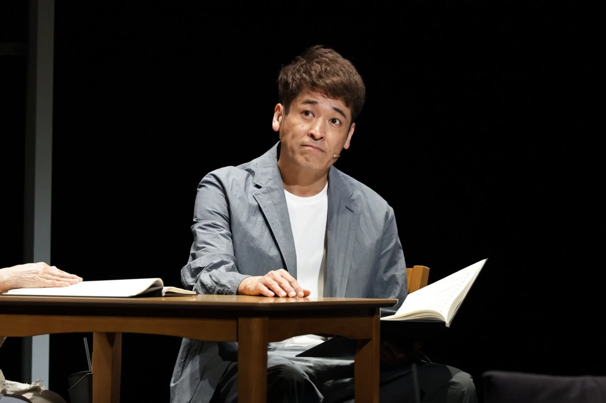 黒い背景の舞台で、一人の男性が木製のテーブルに座り、真剣な表情でカメラを見つめています