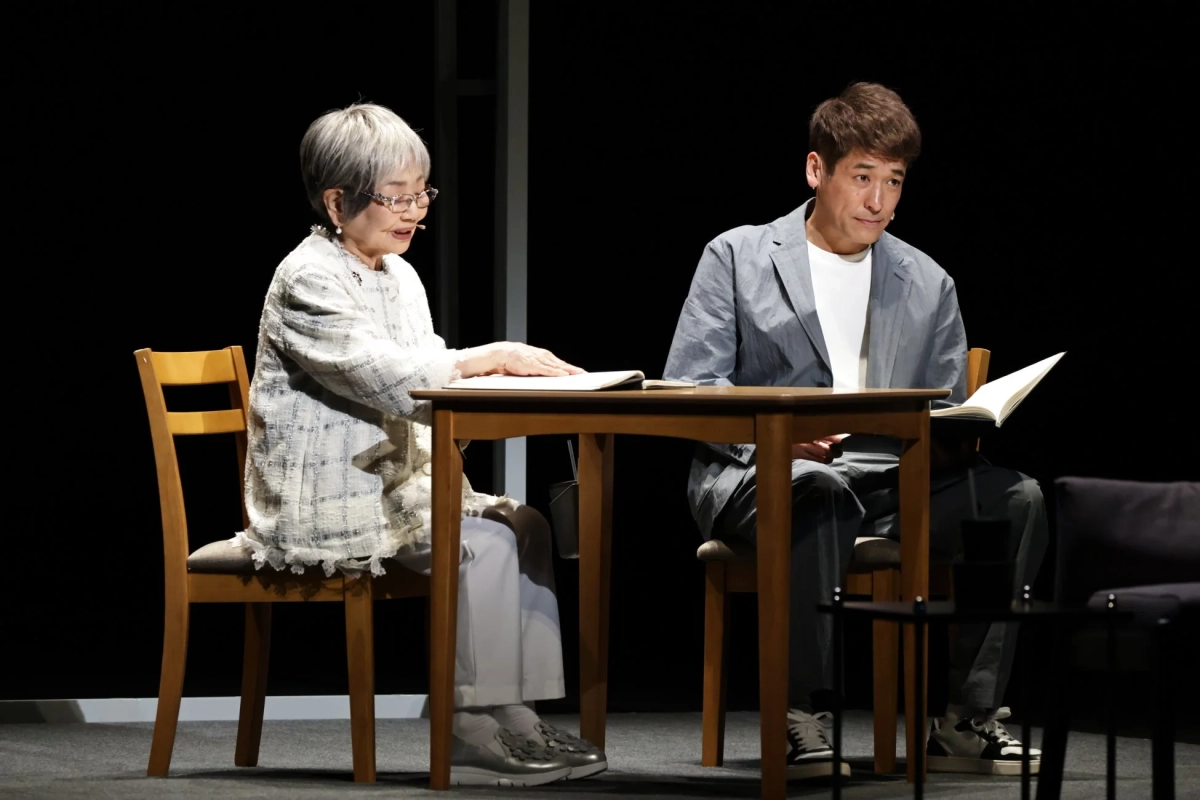 ステージ上で、高齢の女性と中年男性が木製のテーブルを挟んで座り、対談している様子