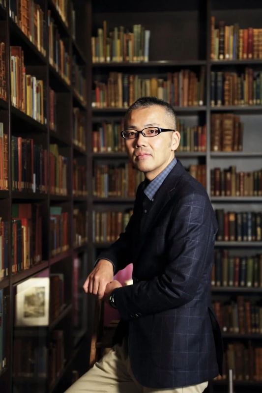 本棚が並ぶ図書館で、メガネをかけた男性がカメラの方を向いて立っています。知的な雰囲気が漂う、落ち着いたポートレートです。