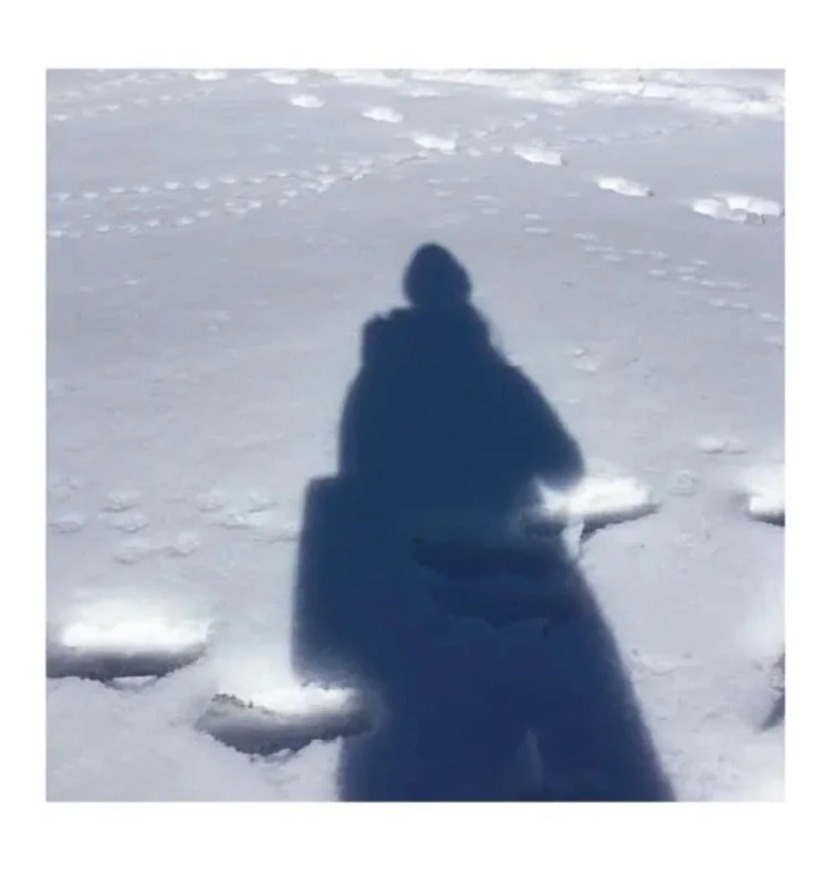 雪原に人の影が長く伸びており、地面には足跡が見られます。冬の晴れた日に屋外で撮影された、静かで広々とした風景の一部を捉えています。