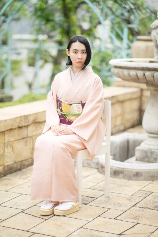薄いピンク色の着物を着た女性が白い椅子に座り、カメラを見つめているポートレート。庭園のような屋外で、背景には緑の木々や石の噴水が見えます。和の落ち着いた雰囲気が特徴です。