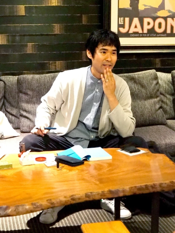 ソファに座り、木製のテーブルを前にして物思いにふけるような男性の姿。背景には「LE JAPON」と書かれたポスターが見え、リラックスした雰囲気の屋内の様子を捉えている。