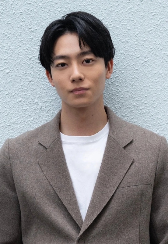 若い東洋人男性が白いTシャツと茶色のジャケットを着用し、水色の壁を背景にカメラを見つめている上半身のポートレート写真です。