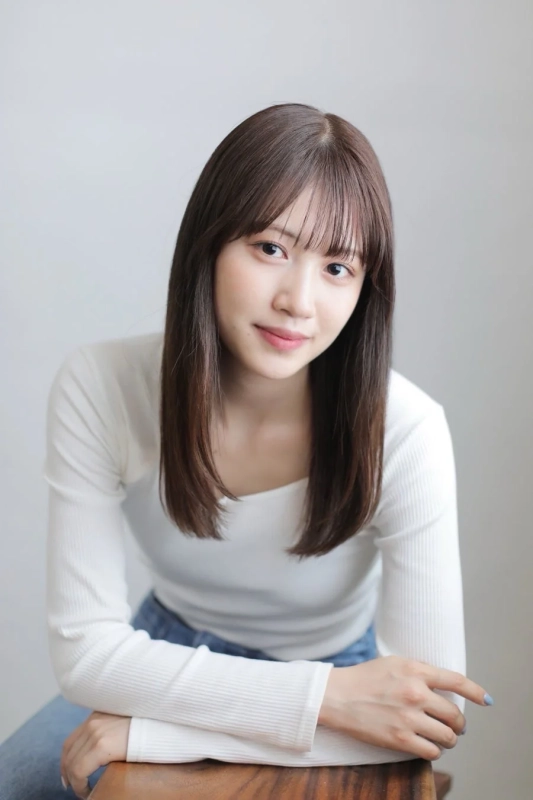 白いリブニットのトップスを着た若い女性が、カメラに向かって優しく微笑んでいます。長めの前髪とストレートのロングヘアが特徴で、明るい背景の前でポーズをとっています。